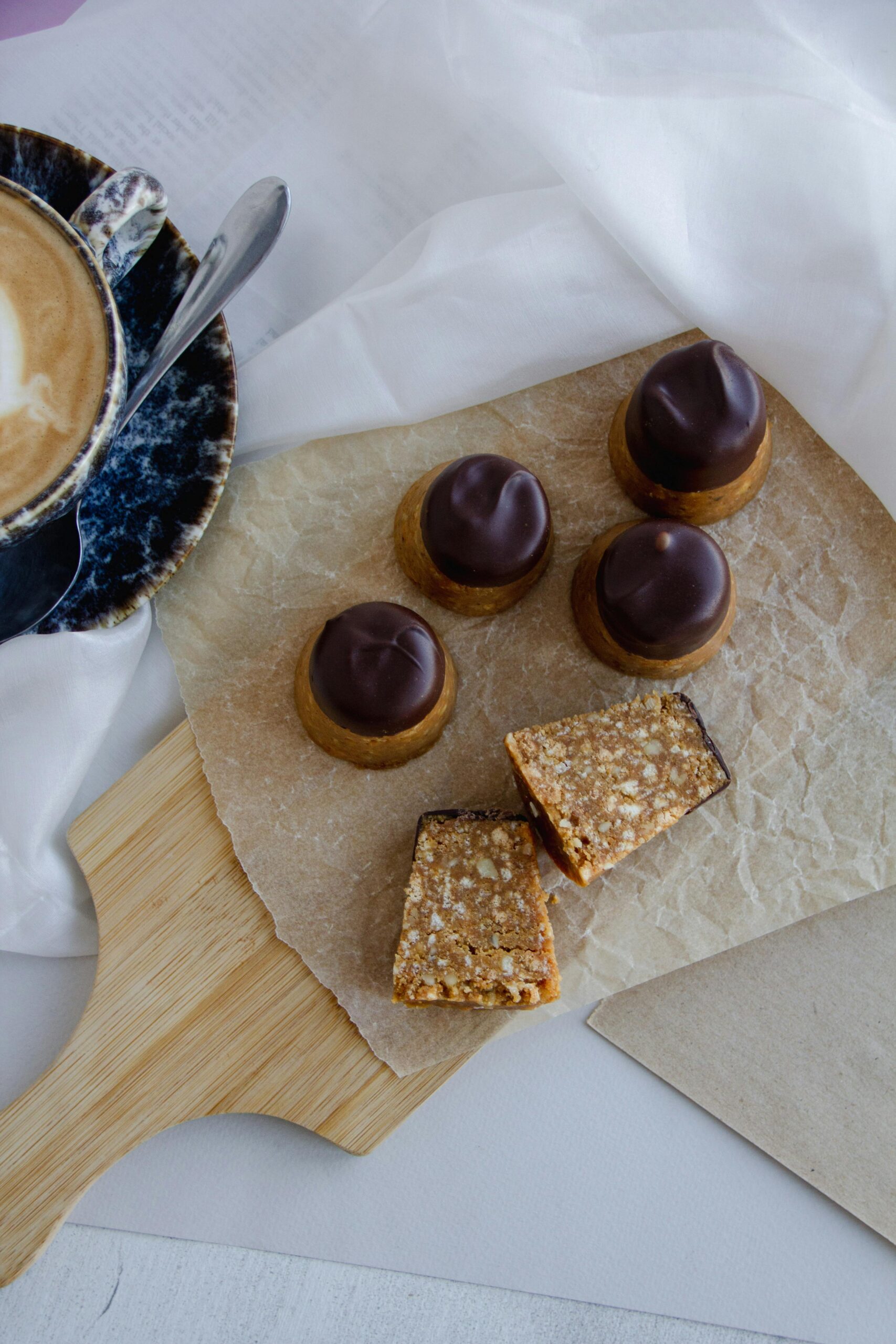 Delightful assortment of chocolate-topped desserts and nut bars on a cutting board with a cappuccino.