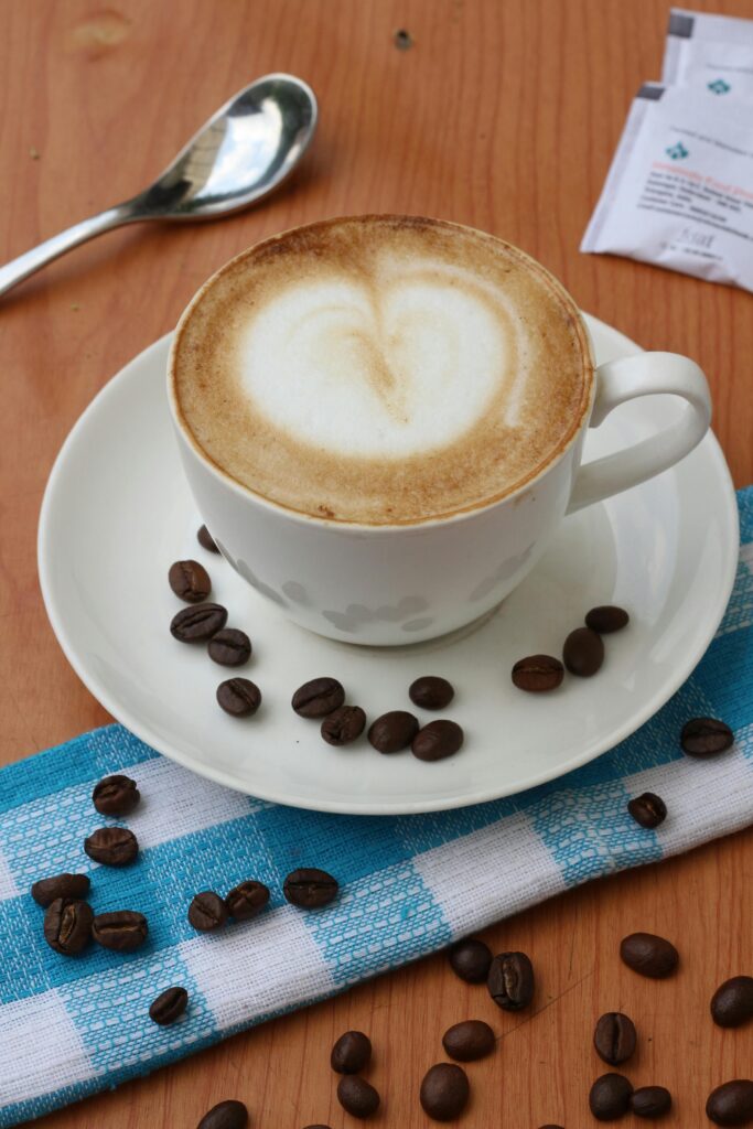 A coffee cup with heart-shaped latte art surrounded by coffee beans on a checkered cloth.